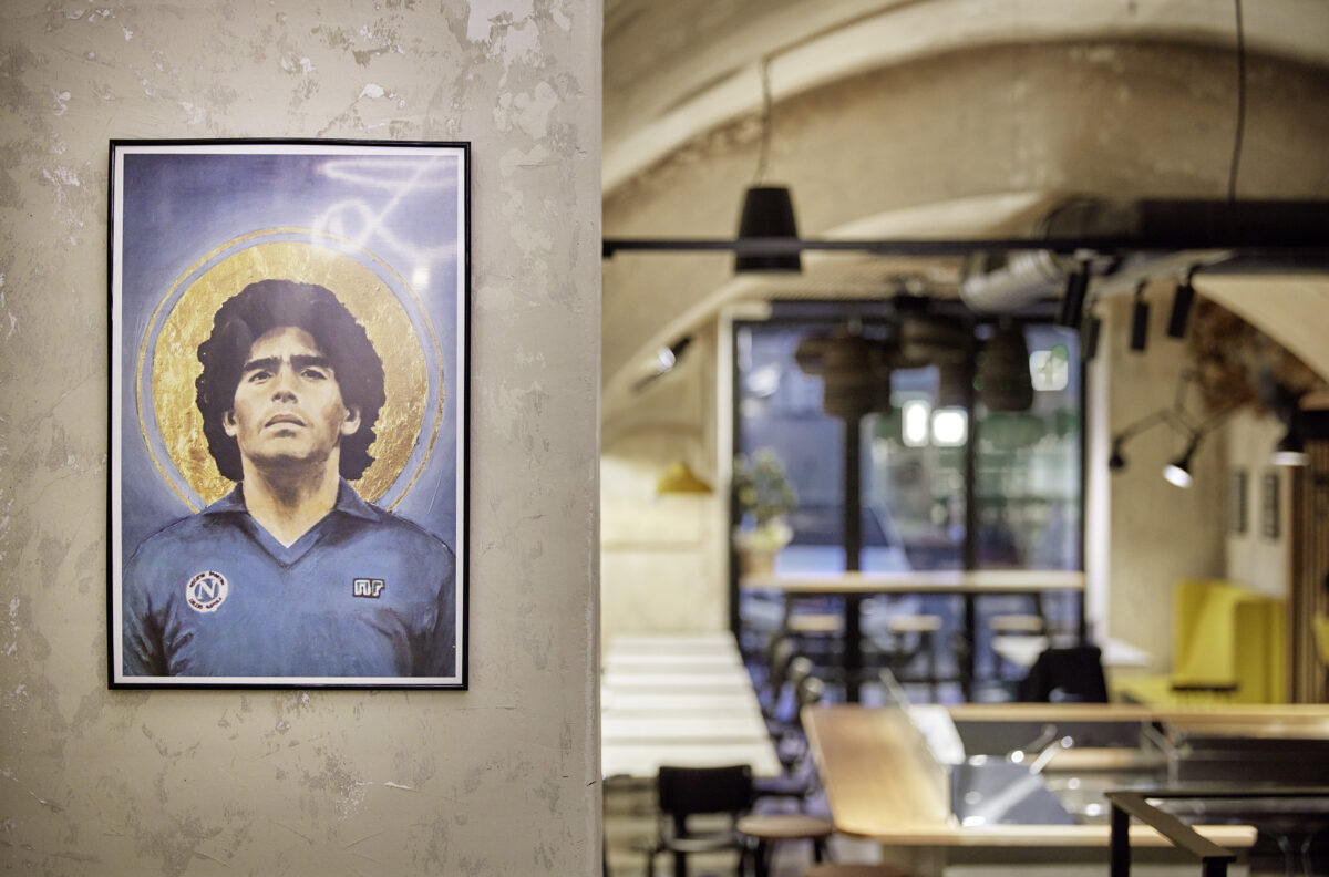 Stimmungsvolles Restaurant-Interieur im De Michele in Vöcklabruck mit einem markanten Wandbild von Diego Maradona auf einer strukturierten Wand, gestaltet von Planbox Innenarchitektur, fotografiert von Roland Wimmer.